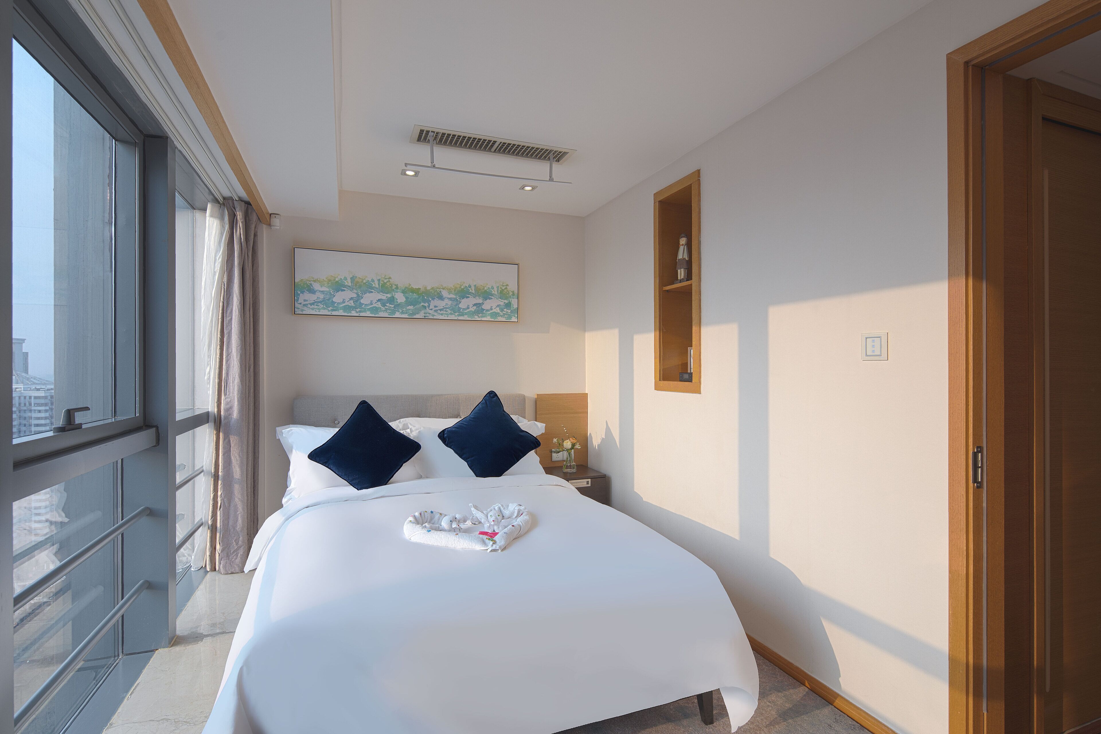 Een hotelkamer met een groot bed, uitzicht op de stad door een raam, een schilderij aan de muur, en een houten plank met een fles.