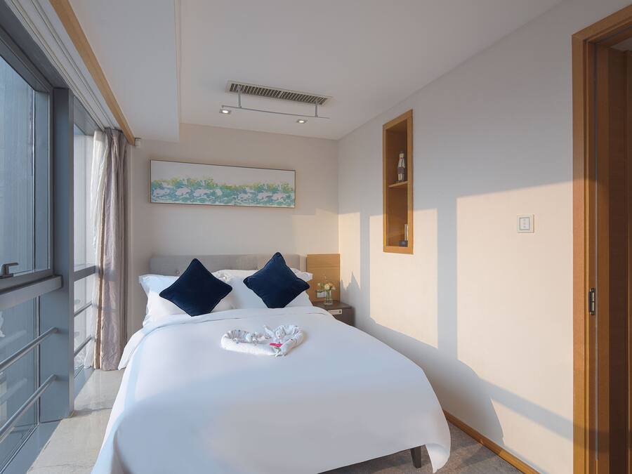 Habitación de hotel con una cama grande, vistas de la ciudad a través de una ventana, un cuadro en la pared y una estantería de madera con una botella.