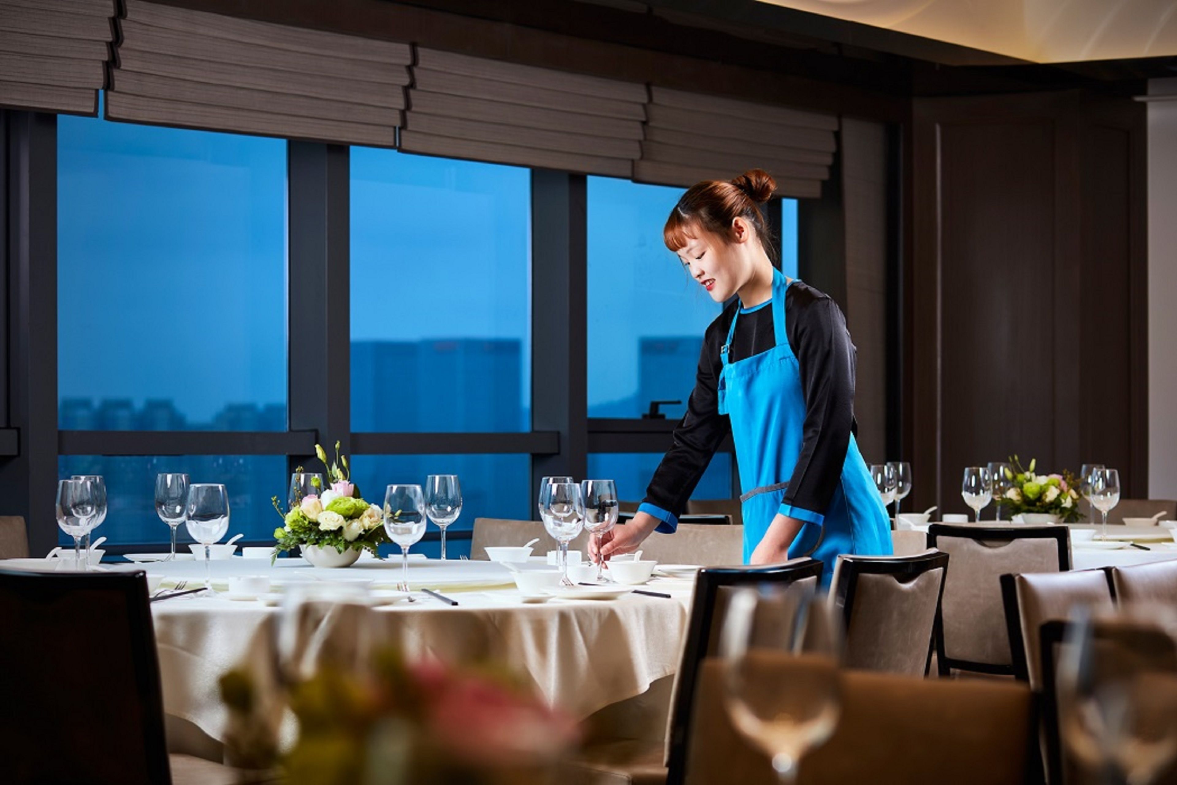 A dining area with tables set for service, a view of the city through large windows, and a person in a blue uniform attending to the tables.