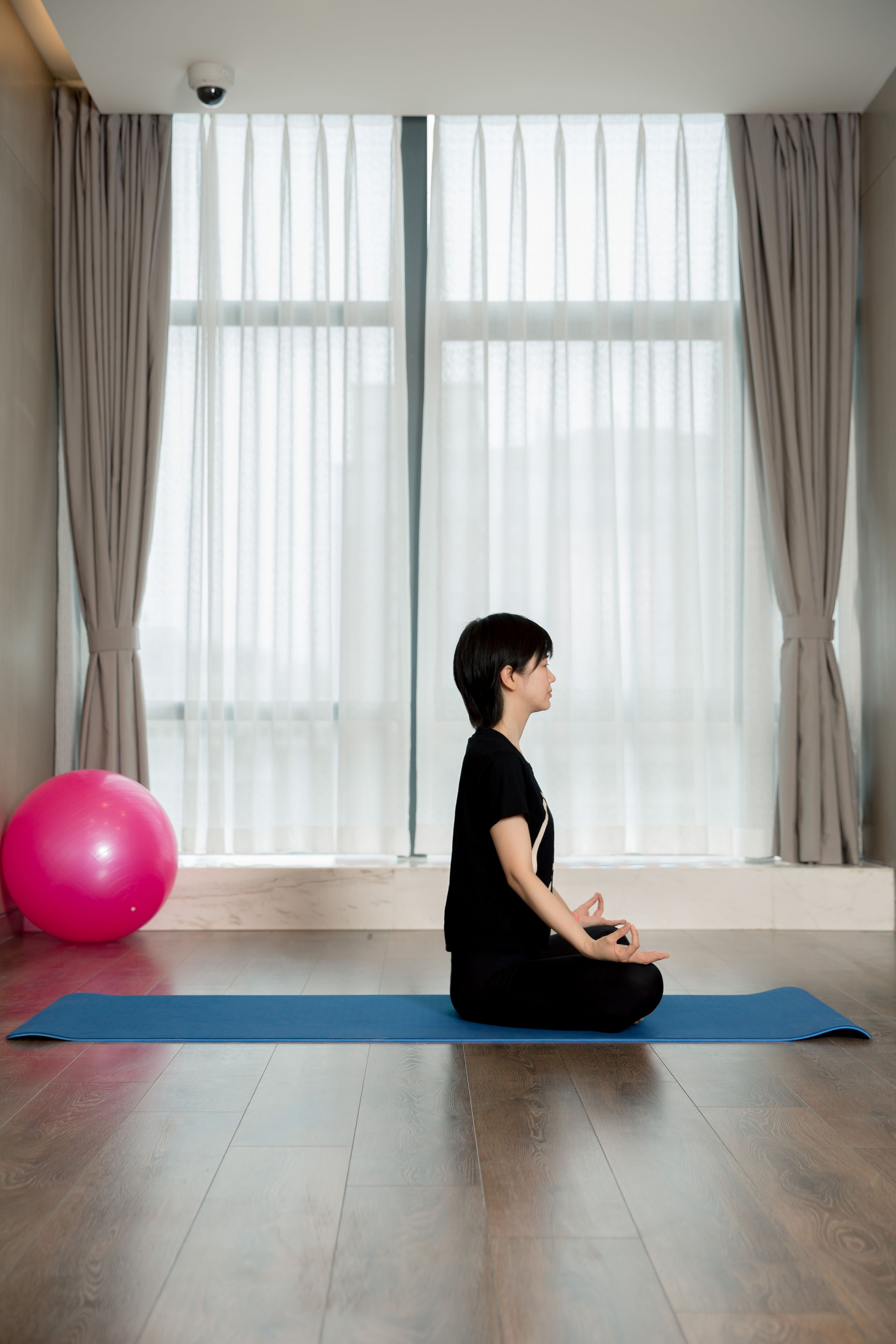 A person sitting on a yoga mat in a room with a large window and curtains.