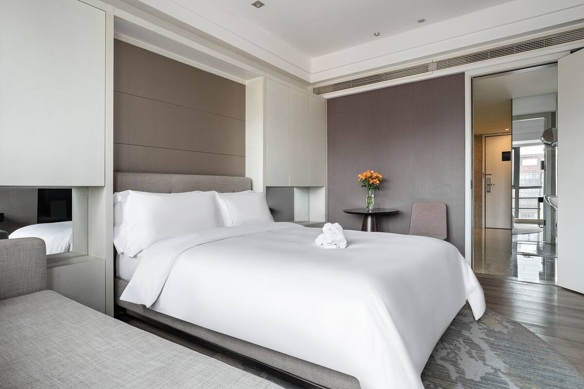 Ein modernes Hotelzimmer mit einem großen Bett, einem Nachttisch mit Blumenvase und Blick auf die Stadt durch das Fenster.