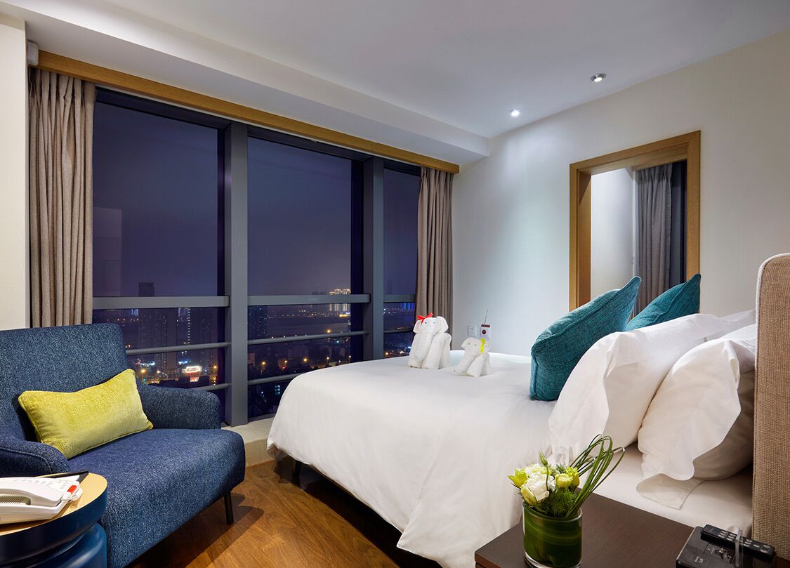A hotel room with a large bed, a sofa, a nightstand, and a view of the city at night.