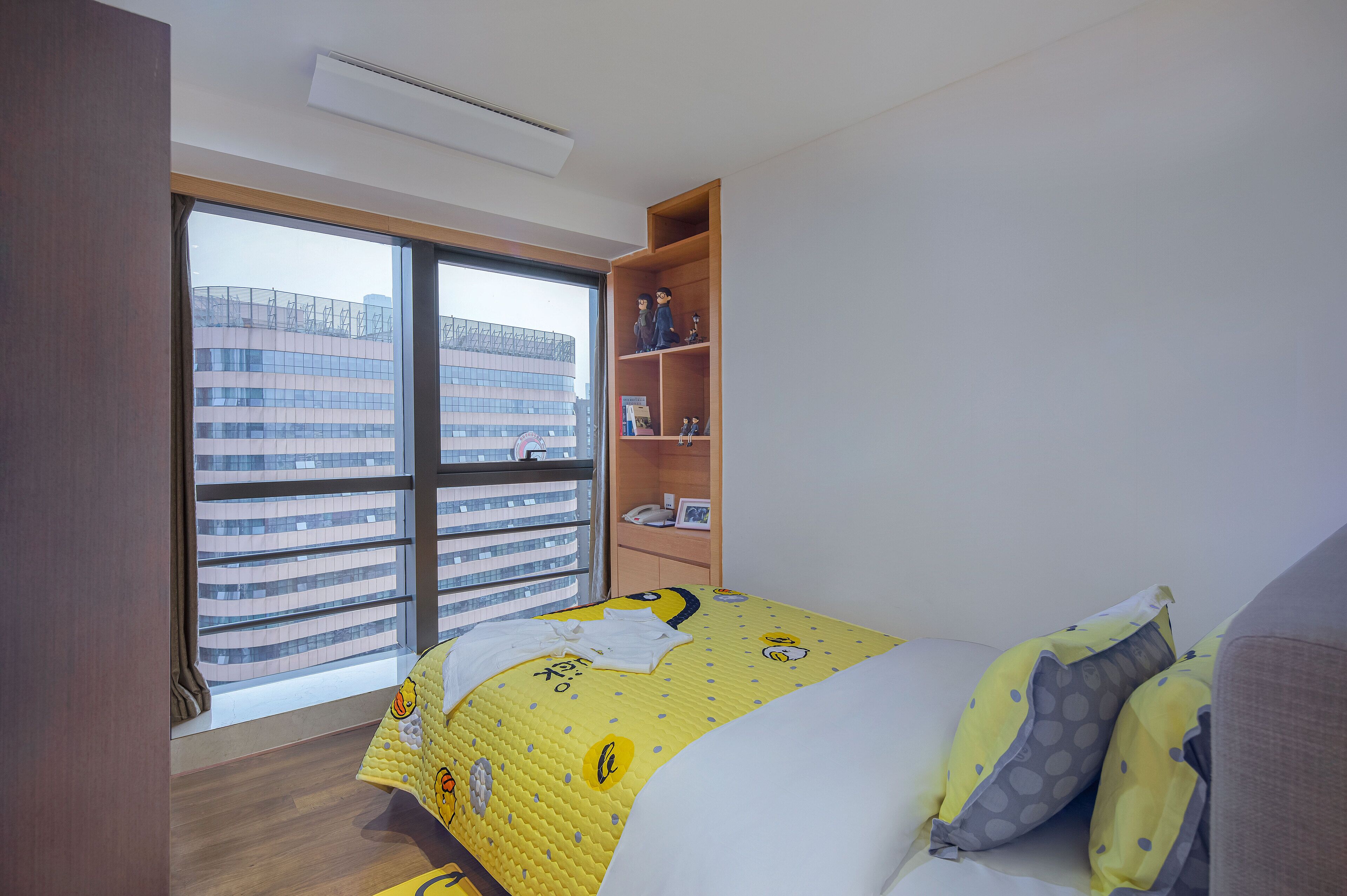 Ein Hotelzimmer mit großem Fenster, einem Bett mit gelber Tagesdecke, einer Holzregaleinrichtung und Blick auf ein modernes Gebäude.
