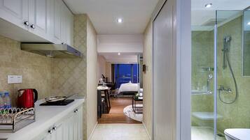 A modern hotel room with a kitchen area, a separate bedroom, and a glass-enclosed shower.