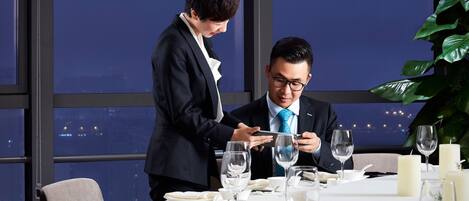 A dining table set for two with a cityscape view, a waiter, and a guest.