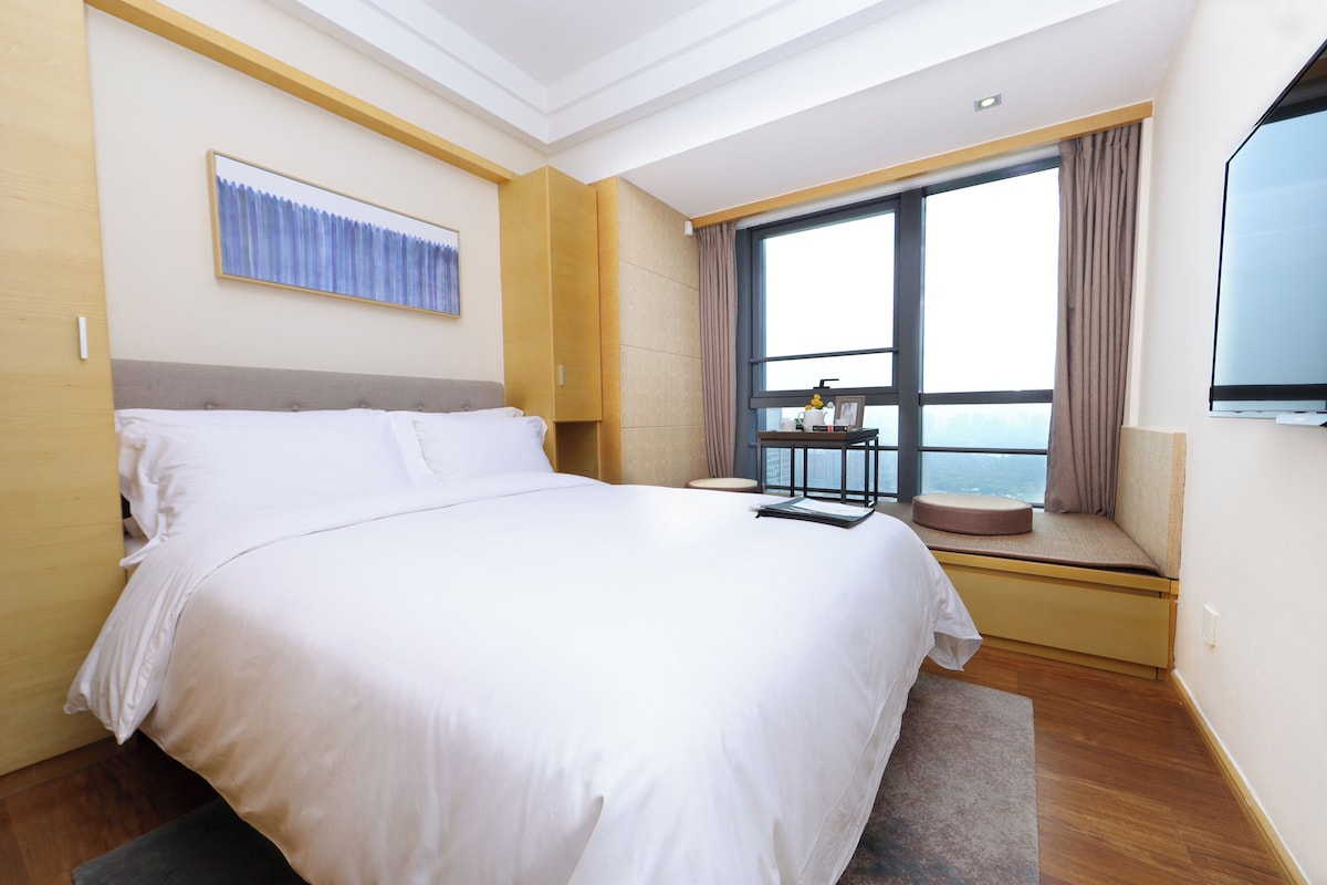 A hotel room with a large bed, a window with a view, a small table with a chair, and a mounted TV.