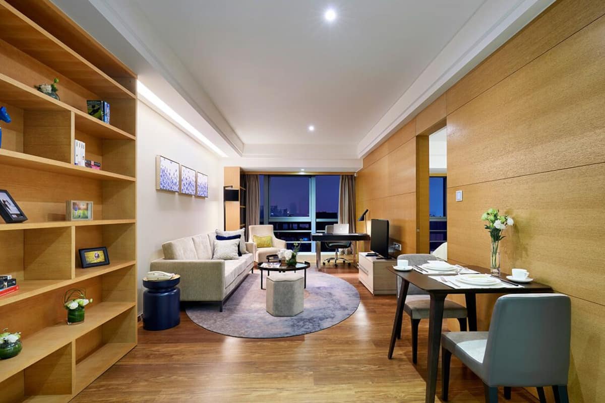 Ein modernes Wohnzimmer mit Sofa, Couchtisch und Essbereich. Ein Bücherregal, ein Fernseher und ein Schreibtisch sind ebenfalls vorhanden. Der Raum verfügt über Holzböden und ein großes Fenster mit Blick auf die Stadt.