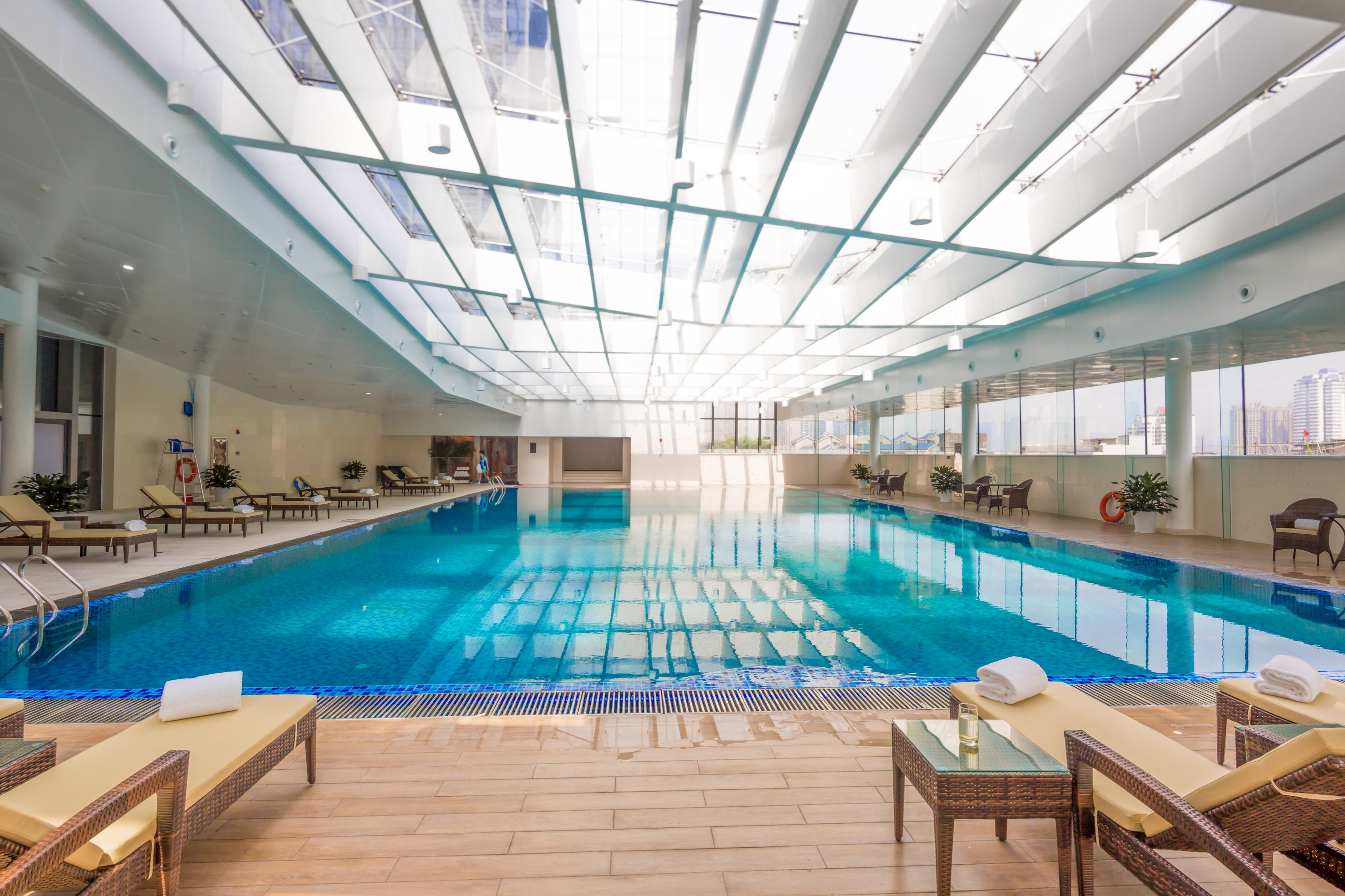 An indoor swimming pool with clear blue water, surrounded by lounge chairs and tables, and large windows offering a view of the city.