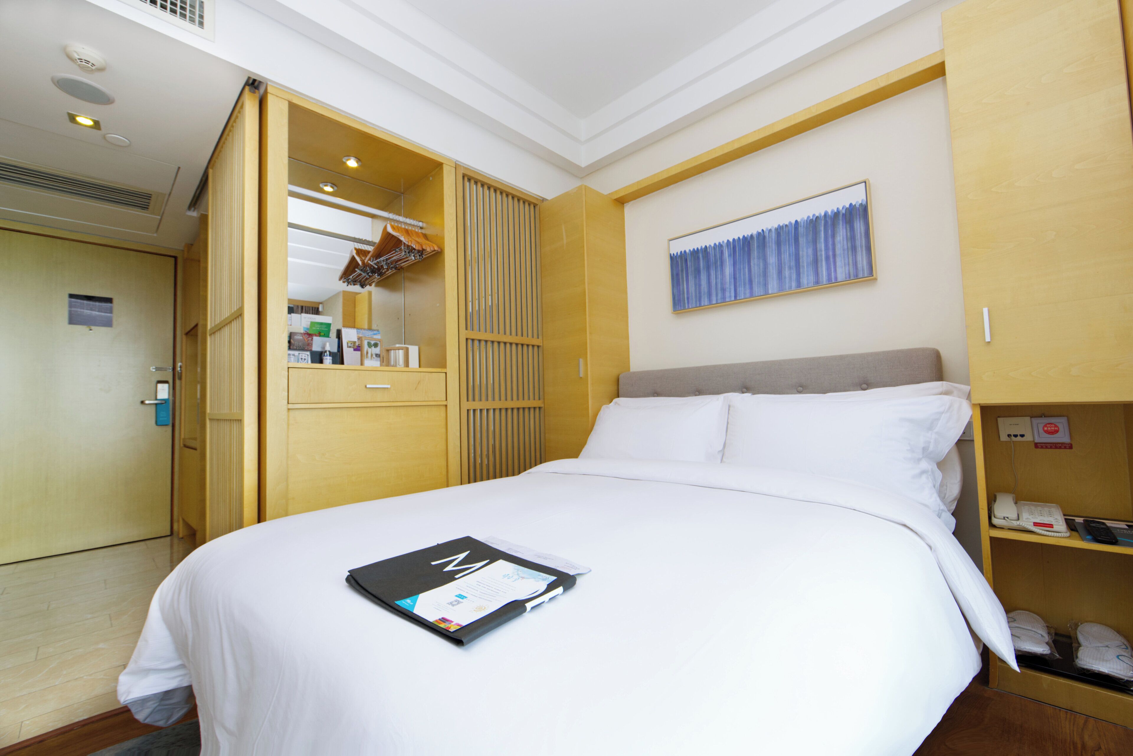 A hotel room with a bed, a wardrobe, a mirror, a painting, and a phone on a shelf.