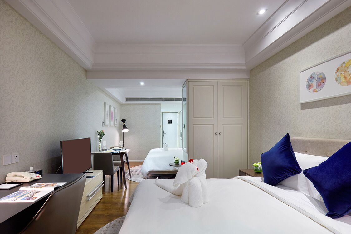 A hotel room with a bed, a desk, a chair, a small table with a lamp, and a mirror.