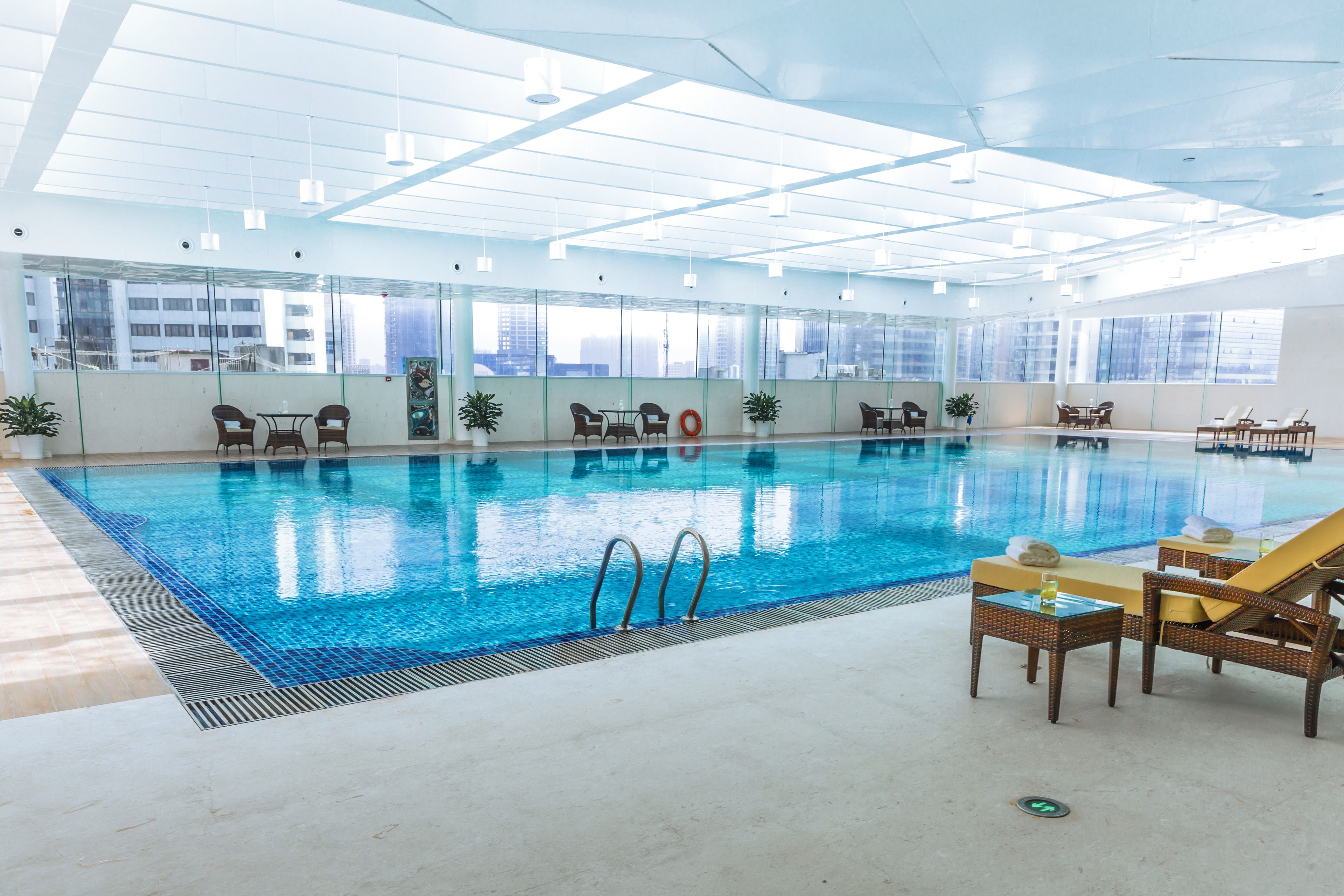An indoor swimming pool with clear glass walls, lounge chairs, and tables.