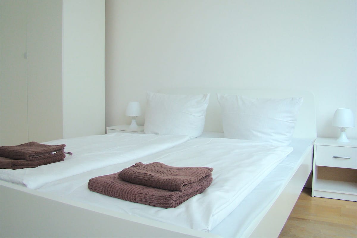 A neatly made bed with white linens and two brown towels, two white pillows, and two bedside lamps on white nightstands.