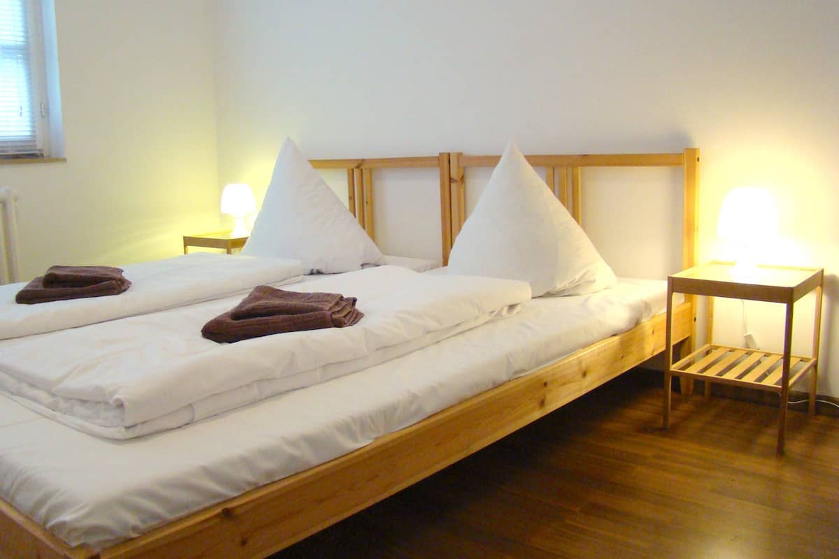 A double bed with white linens, two pillows, and a wooden headboard. There are two bedside tables with lamps, and a brown towel is folded on the bed.