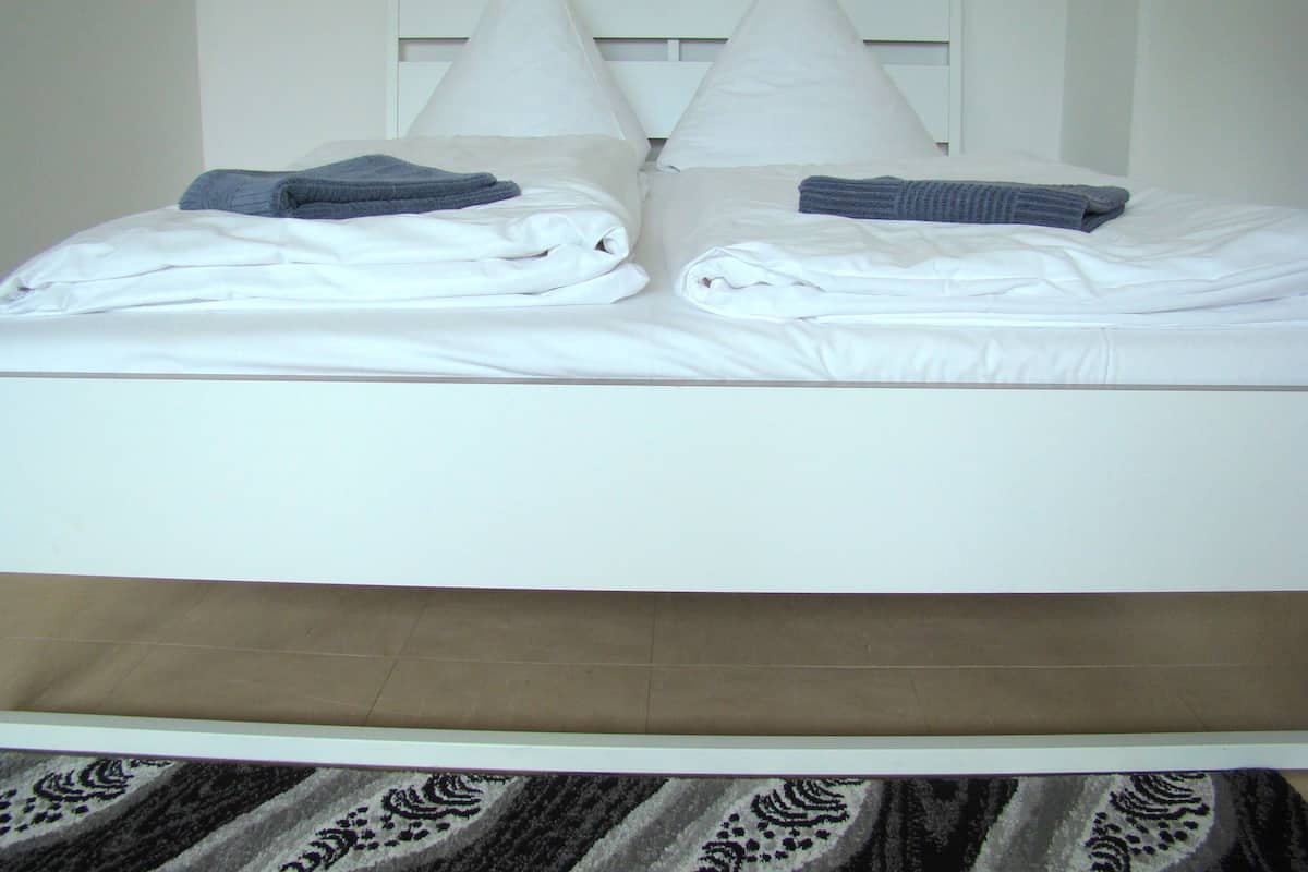 A neatly made bed with white linens and two pillows, a folded towel, and a patterned rug at the foot of the bed.