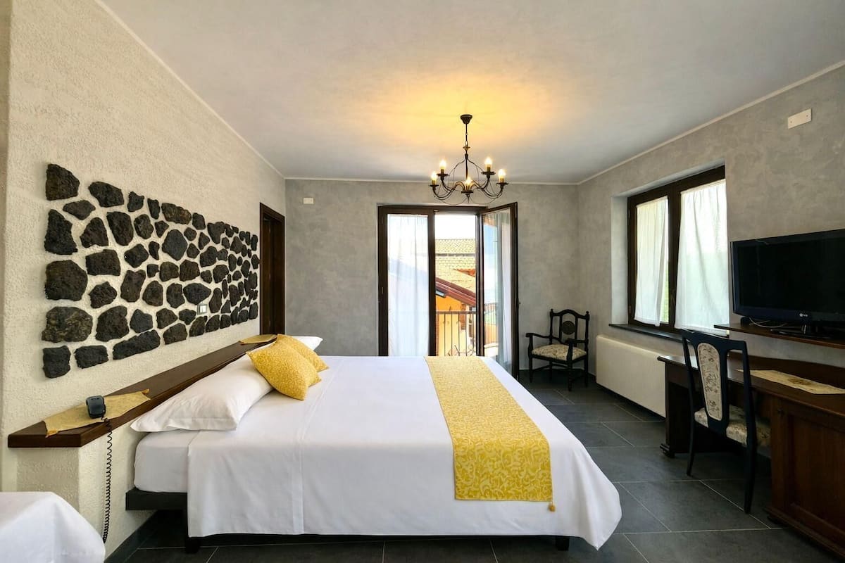 A hotel room with a stone wall, a bed with white bedding and a yellow throw, a desk with a chair, a television, and a balcony with a view.