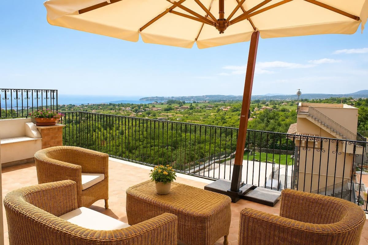 A balcony with wicker furniture, a large umbrella, and a view of a green landscape.