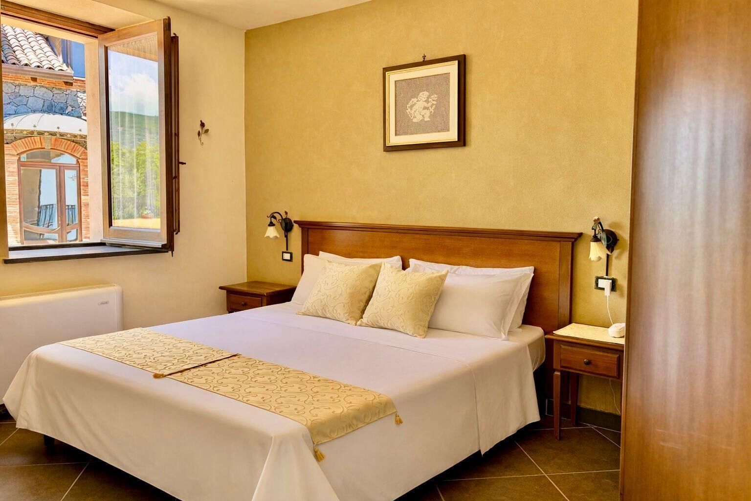 A hotel room with a bed, two bedside tables, a nightstand, a window with a view, and a framed picture on the wall.