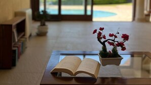 Un libro abierto sobre una mesa con una planta en maceta, una estantería y vistas a través de amplias puertas de cristal.