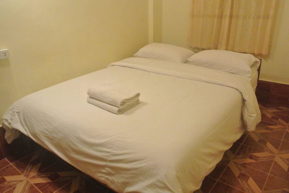 A neatly made bed with a folded towel, a plain wall, and a patterned floor.