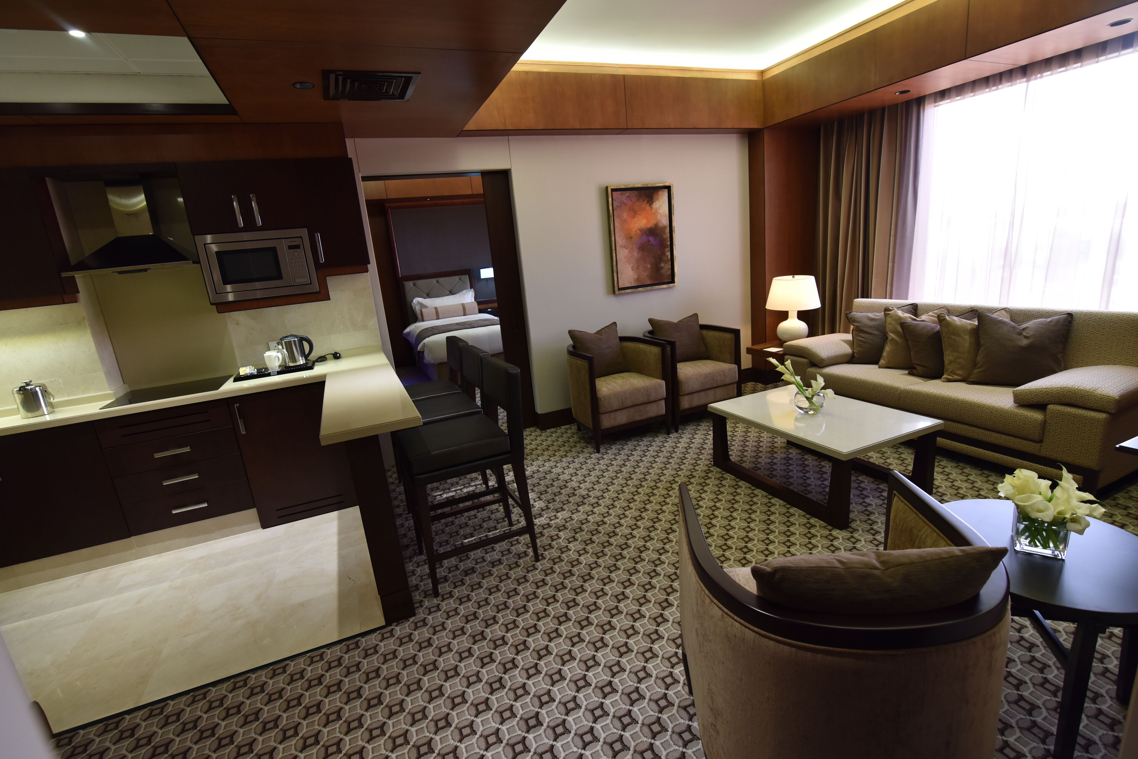 A hotel room with a kitchenette, a dining area, a living room with a sofa and armchairs, and a large window with curtains.