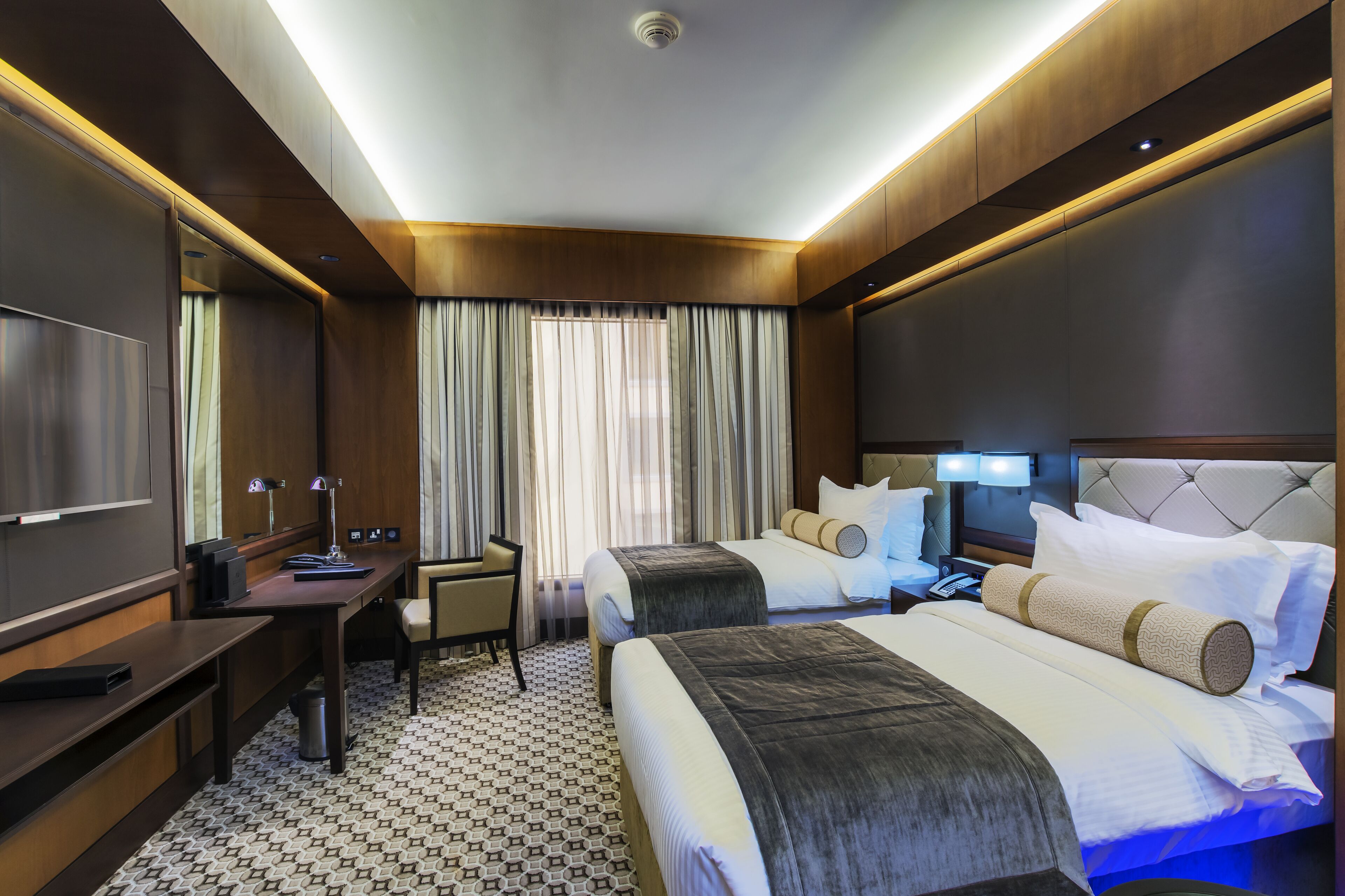 A hotel room with two beds, a desk, a chair, a TV, and a window with curtains.