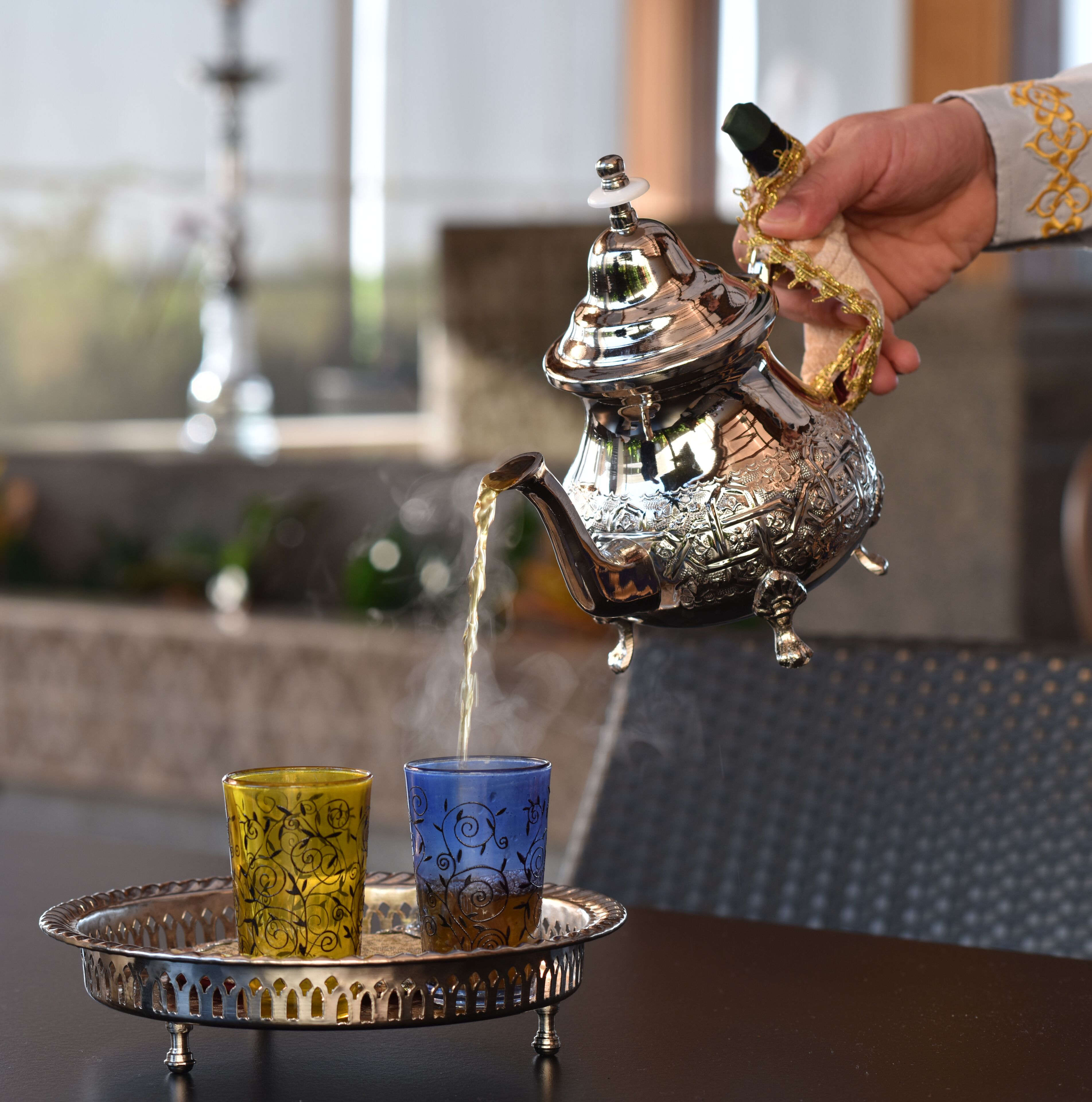 A hand pouring tea from a silver teapot into two glasses on a tray.