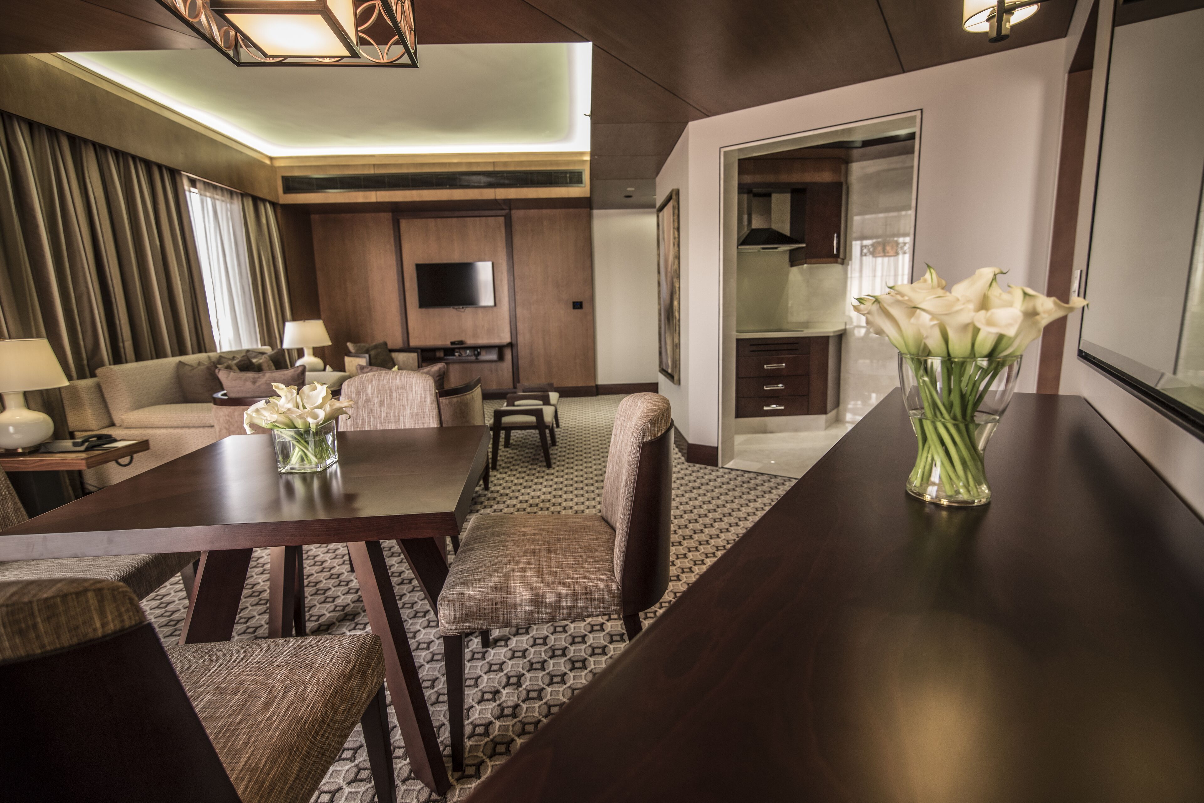 A modern hotel room with a dining area, a sofa set, a flat-screen TV, and a vase of white flowers on the dining table.