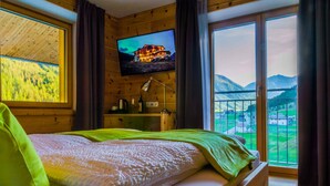 Une chambre à coucher avec un lit, un téléviseur fixé au mur et une vue sur un paysage montagneux, visible à travers une grande fenêtre.