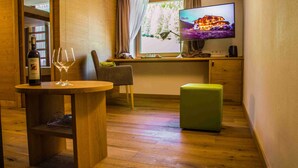 Une pièce chaleureuse avec un plancher en bois, une petite table ronde sur laquelle se trouvent une bouteille et des verres, un pouf vert, une chaise et un téléviseur qui diffuse une image d’une maison.
