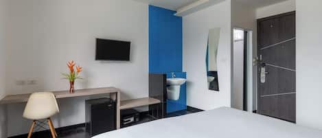 A modern hotel room with a bed, a desk, a chair, a TV, a sink, and a door.