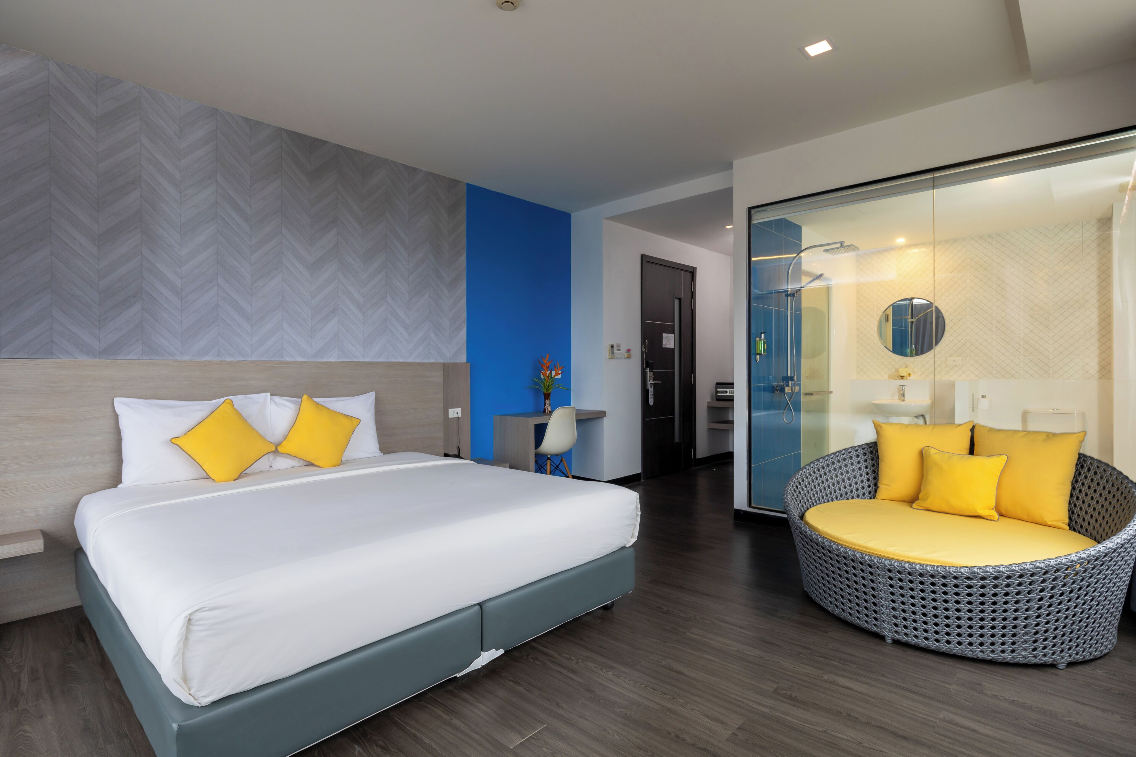A modern hotel room with a large bed, a round wicker sofa, and a glass shower enclosure.