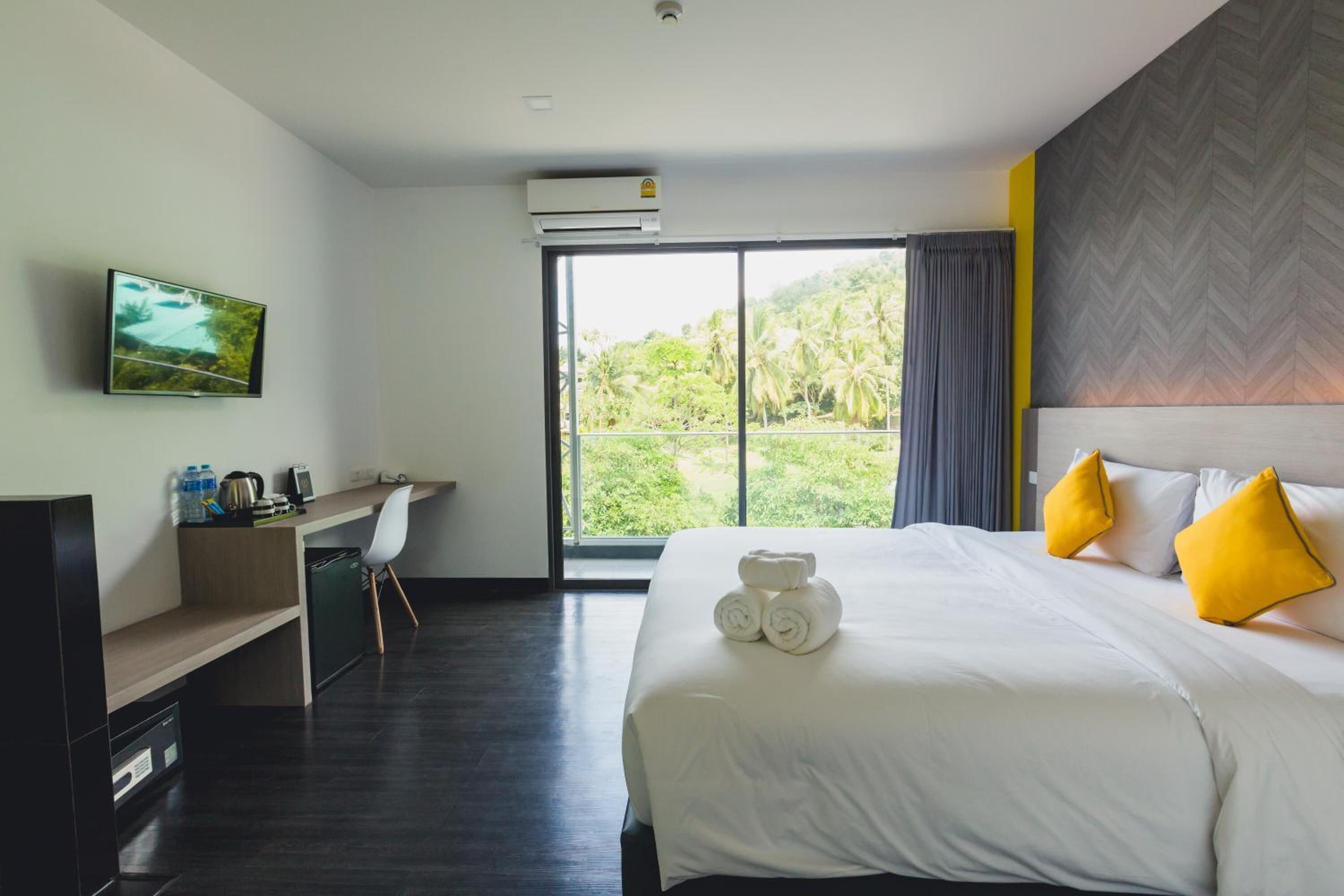 Habitación de hotel con cama, escritorio, silla, televisor y ventana grande con vista a la vegetación.