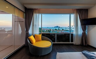 Habitación de hotel con un ventanal grande que ofrece vistas al mar y a edificios. Hay una silla de mimbre con cojines amarillos y una cama con ropa blanca.