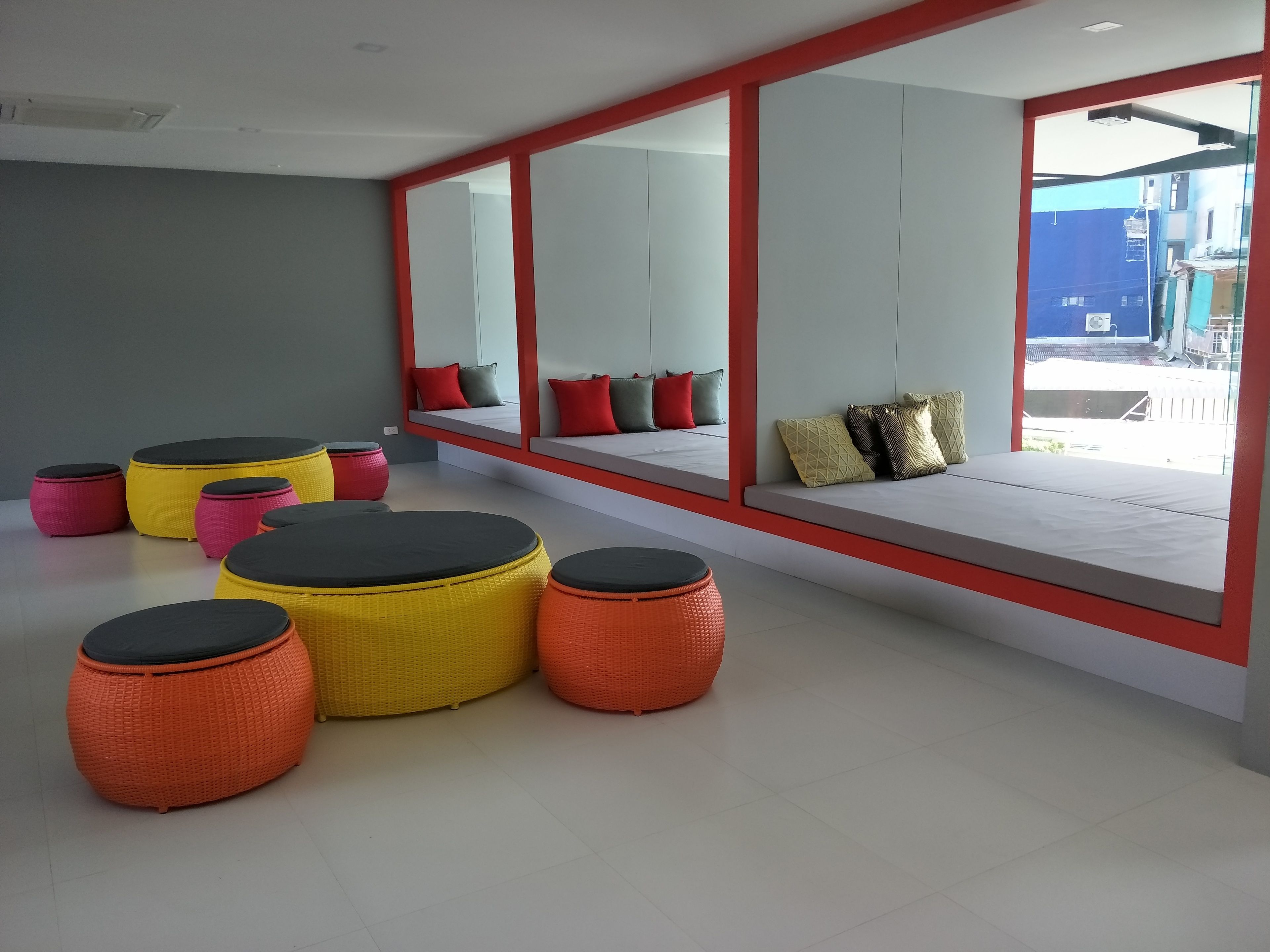 Ein modernes Interieur mit einer Sitzecke, ausgestattet mit runden, gewebten Poufs in verschiedenen Farben, einem großen Fenster mit Ausblick und einer roten Wand.