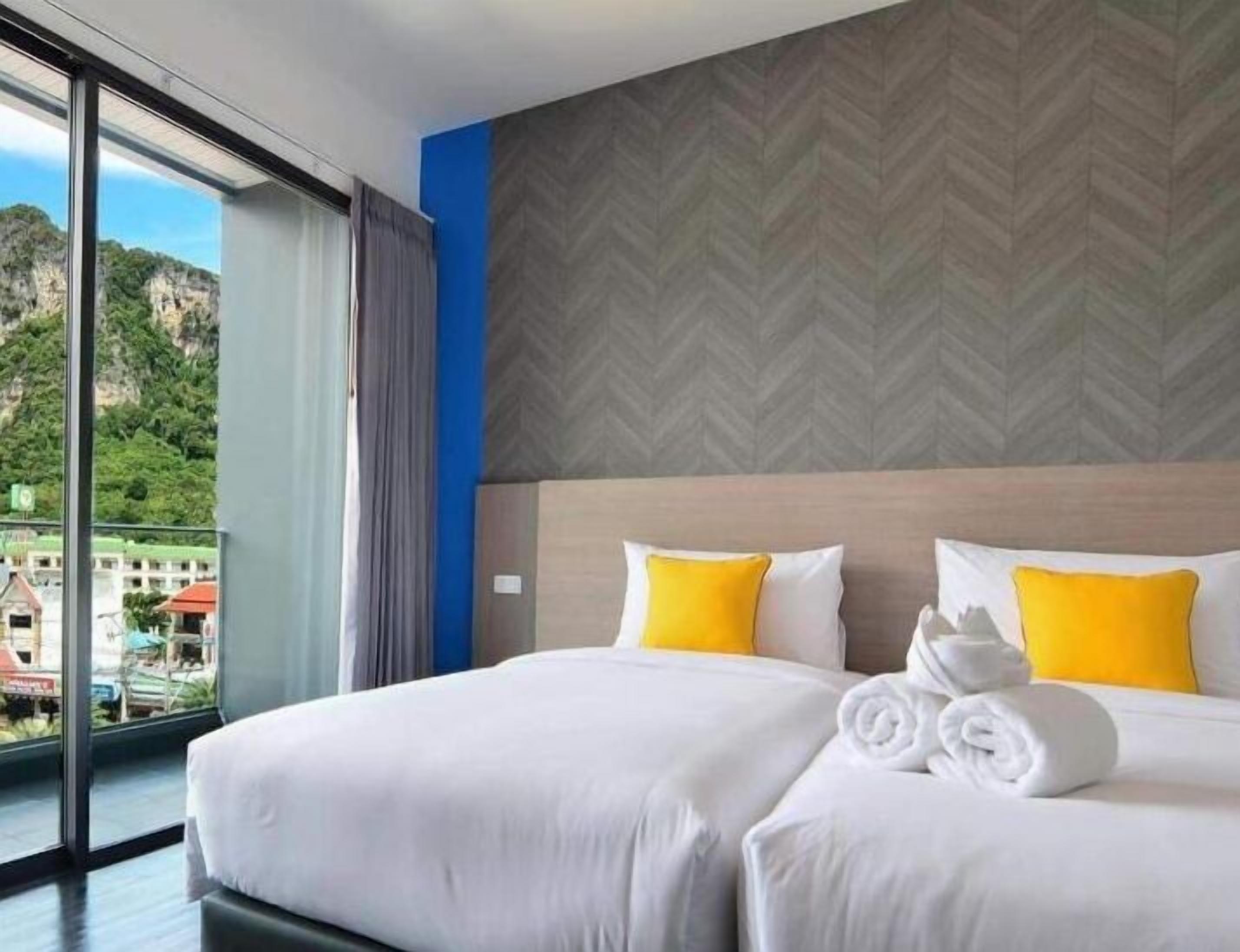 Une chambre d’hôtel avec une grande fenêtre, un lit recouvert d’une literie blanche et agrémenté de deux oreillers jaunes, et un mur orné d’un motif en chevrons.