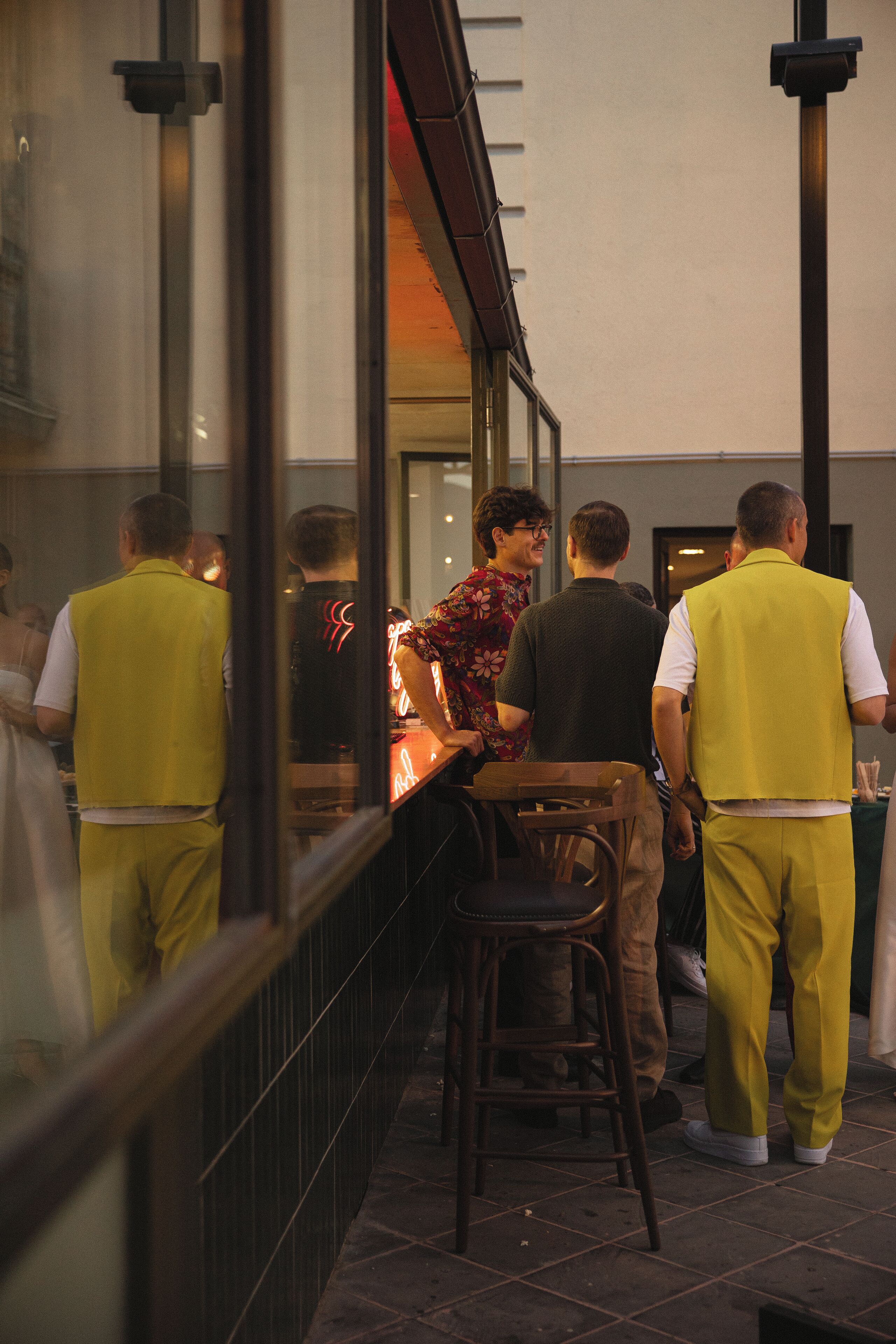 A group of people standing outside a building with a glass window, a chair, and a table.