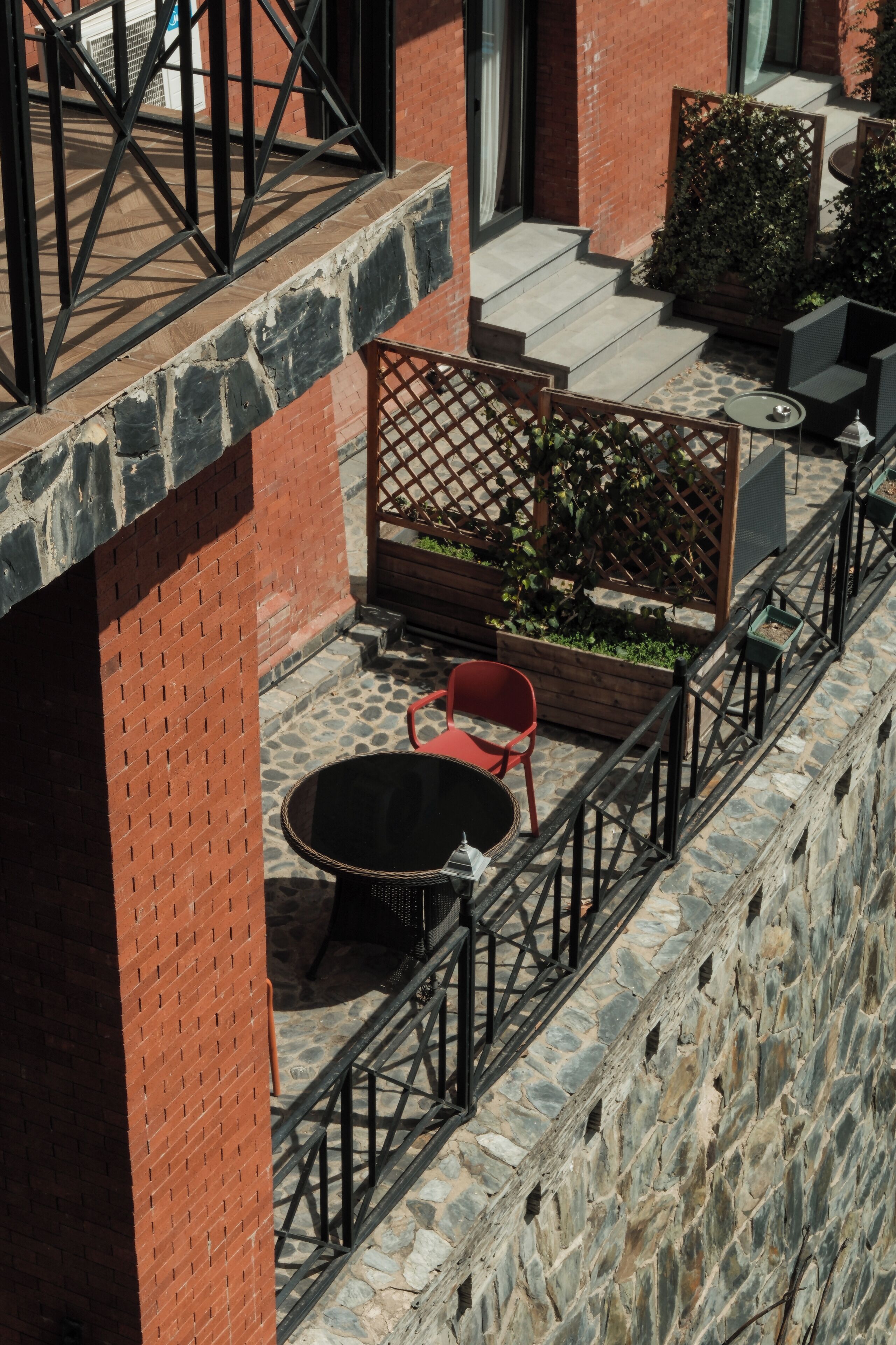 A courtyard with a stone wall, a brick building, a wooden fence, a round table, and a red chair.