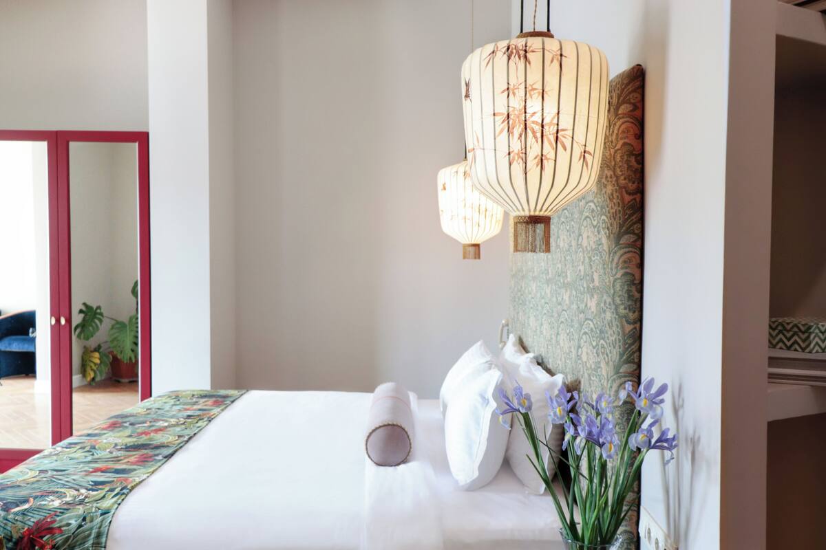 A hotel room with a bed, a hanging light fixture, a potted plant, and a view of the outside.