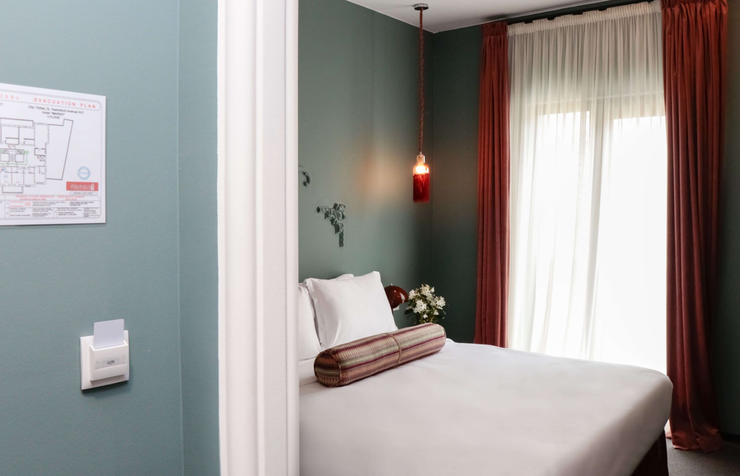 A hotel room with a bed, a wall-mounted emergency exit plan, a wall-mounted paper dispenser, and a hanging pendant light.
