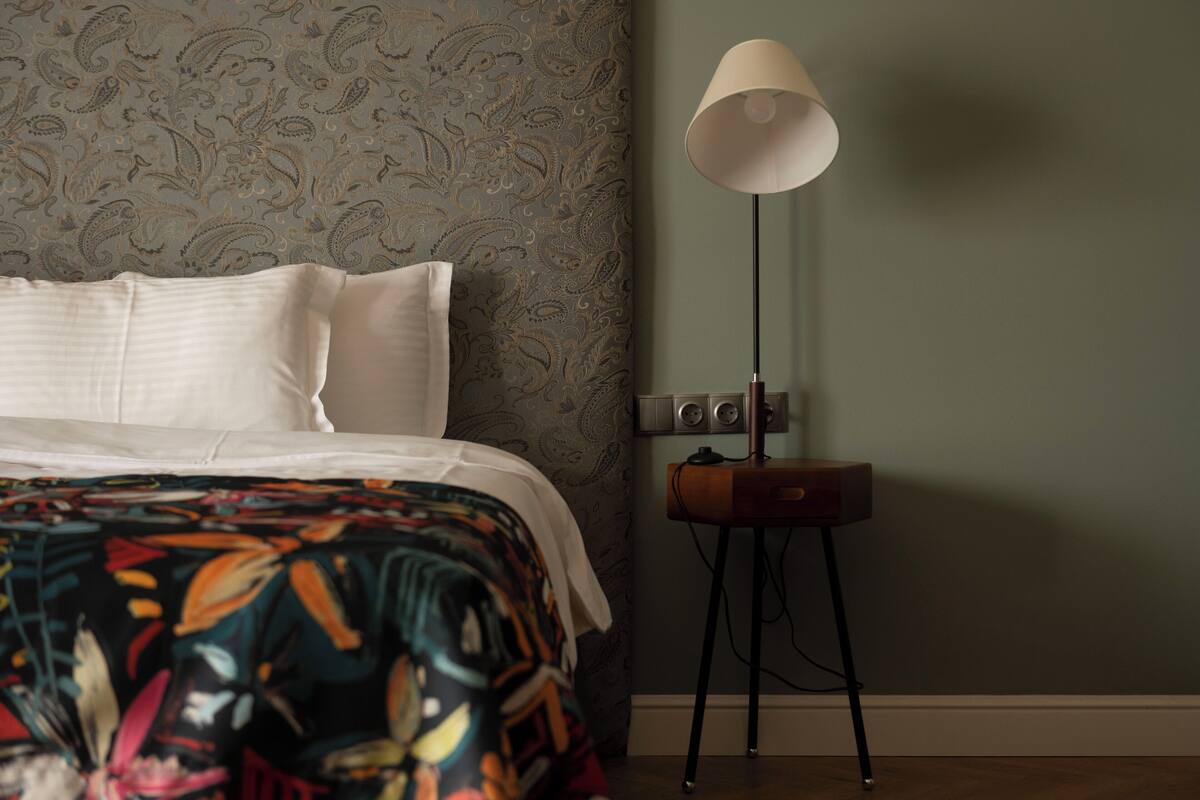 A bedroom with a bed, patterned wallpaper, a standing lamp, and a small wooden side table.