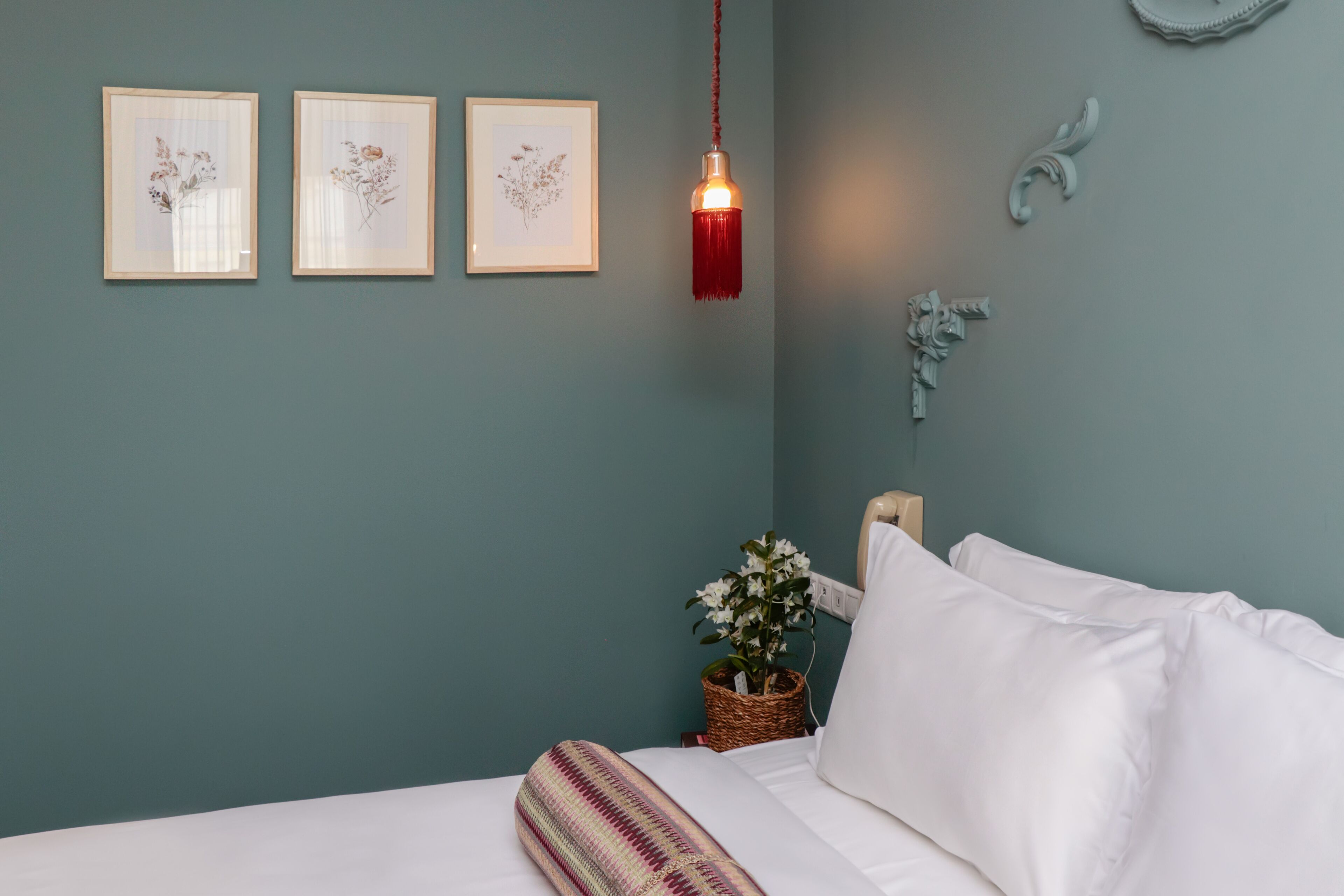 A neatly made bed with white linens, a hanging red tassel light fixture, and framed botanical prints on the wall.
