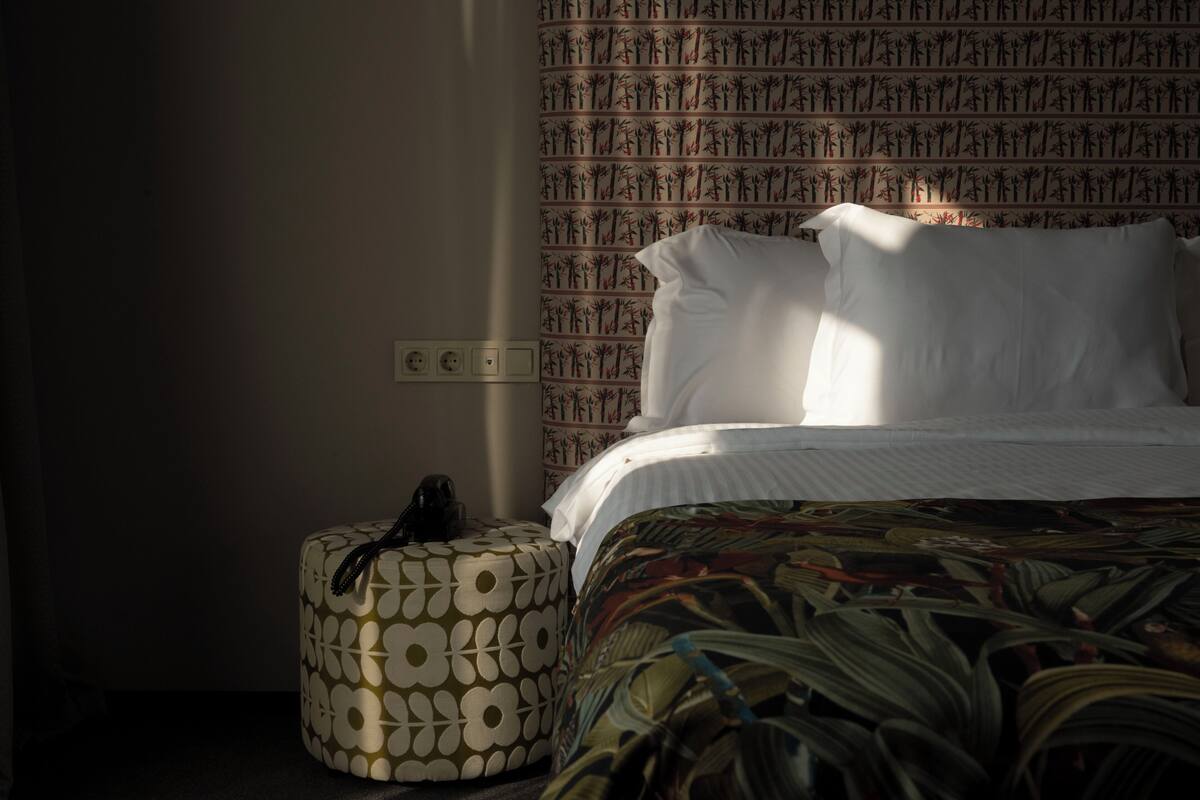 A hotel room with a bed, a patterned ottoman, and a wall with a tree pattern.