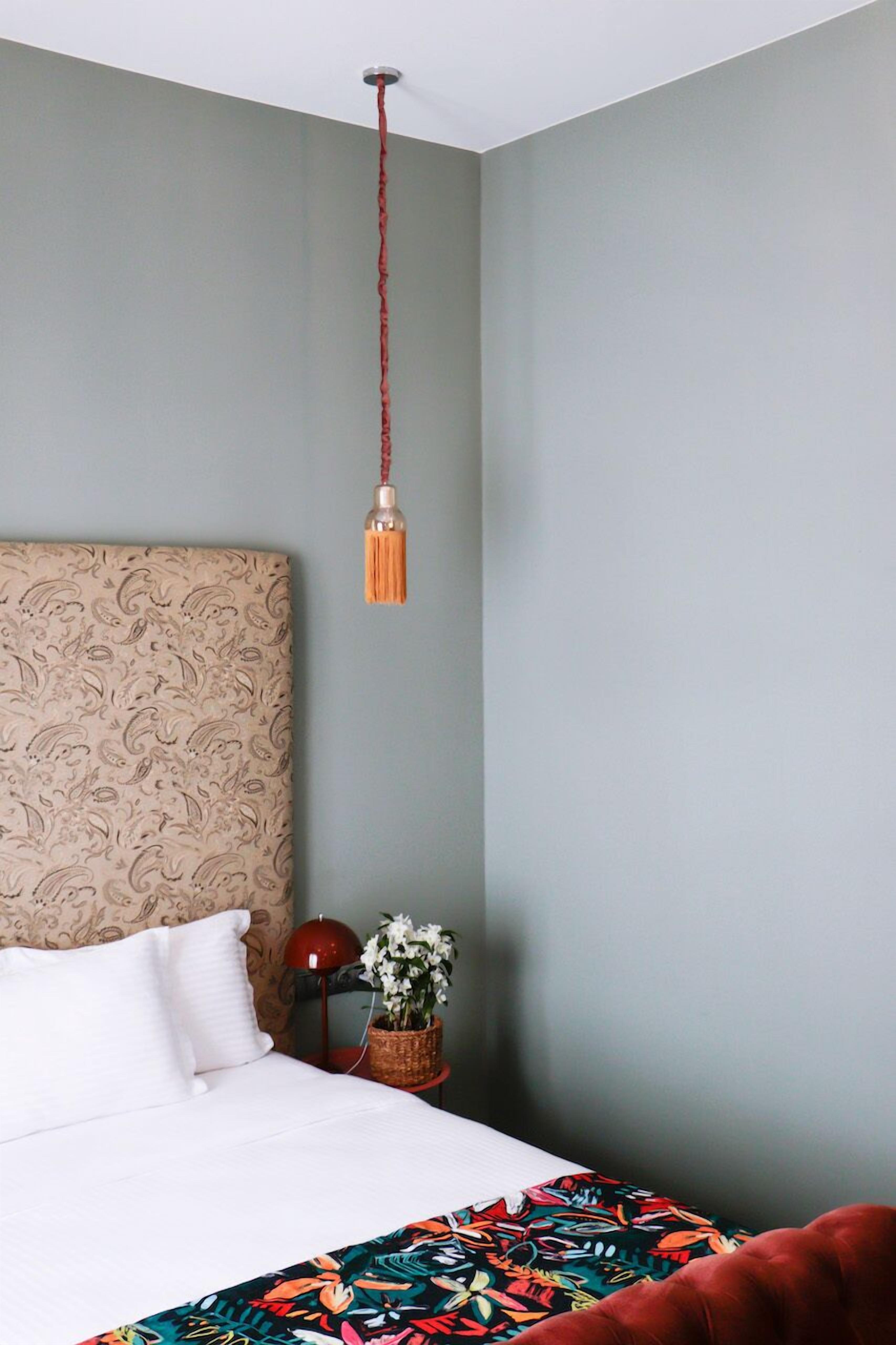 A bedroom with a patterned headboard, a red lamp, a potted plant, and a colorful throw blanket.