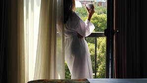 A person in a white robe standing by a window with sheer curtains, holding a cup. - Margot Old Tbilisi (Tbilisi)