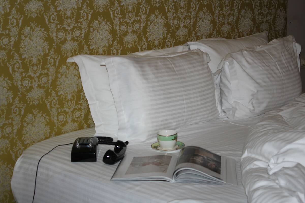 A neatly made bed with white linens, a book, a cup, and a telephone on a bedside table.