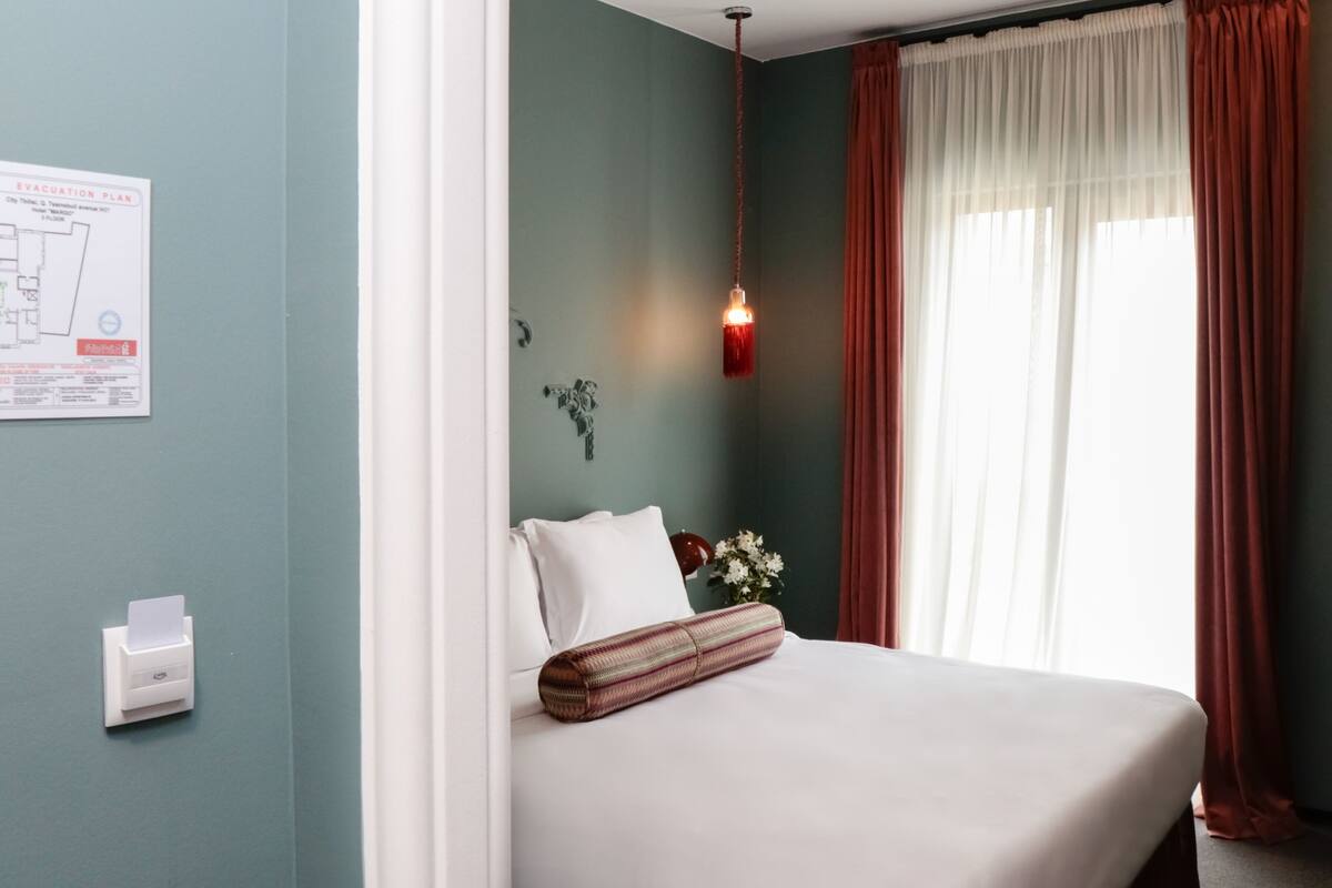 A hotel room with a bed, a wall-mounted emergency exit plan, a hanging light fixture, and a window with curtains.