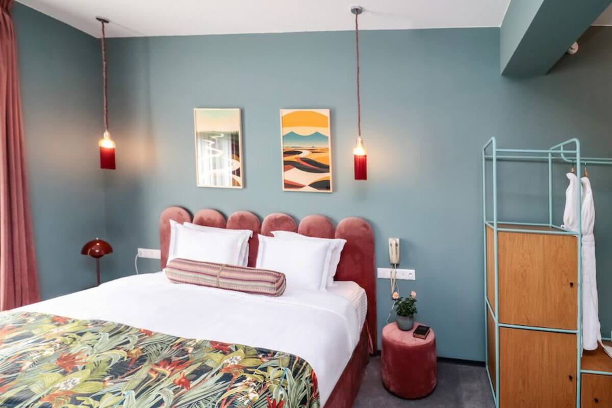 A bedroom with a bed, two wall art pieces, a red lamp, a wooden wardrobe, and a red stool.
