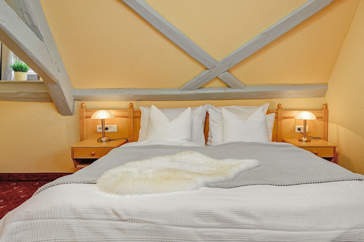 A bedroom with a bed, two bedside tables, two lamps, and a window with a plant.
