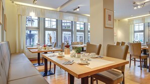 A dining area with wooden tables and chairs, large windows with curtains, and a view of the street outside. - Hotel Mühlhäuser Hof (Mühlhausen/Thüringen)