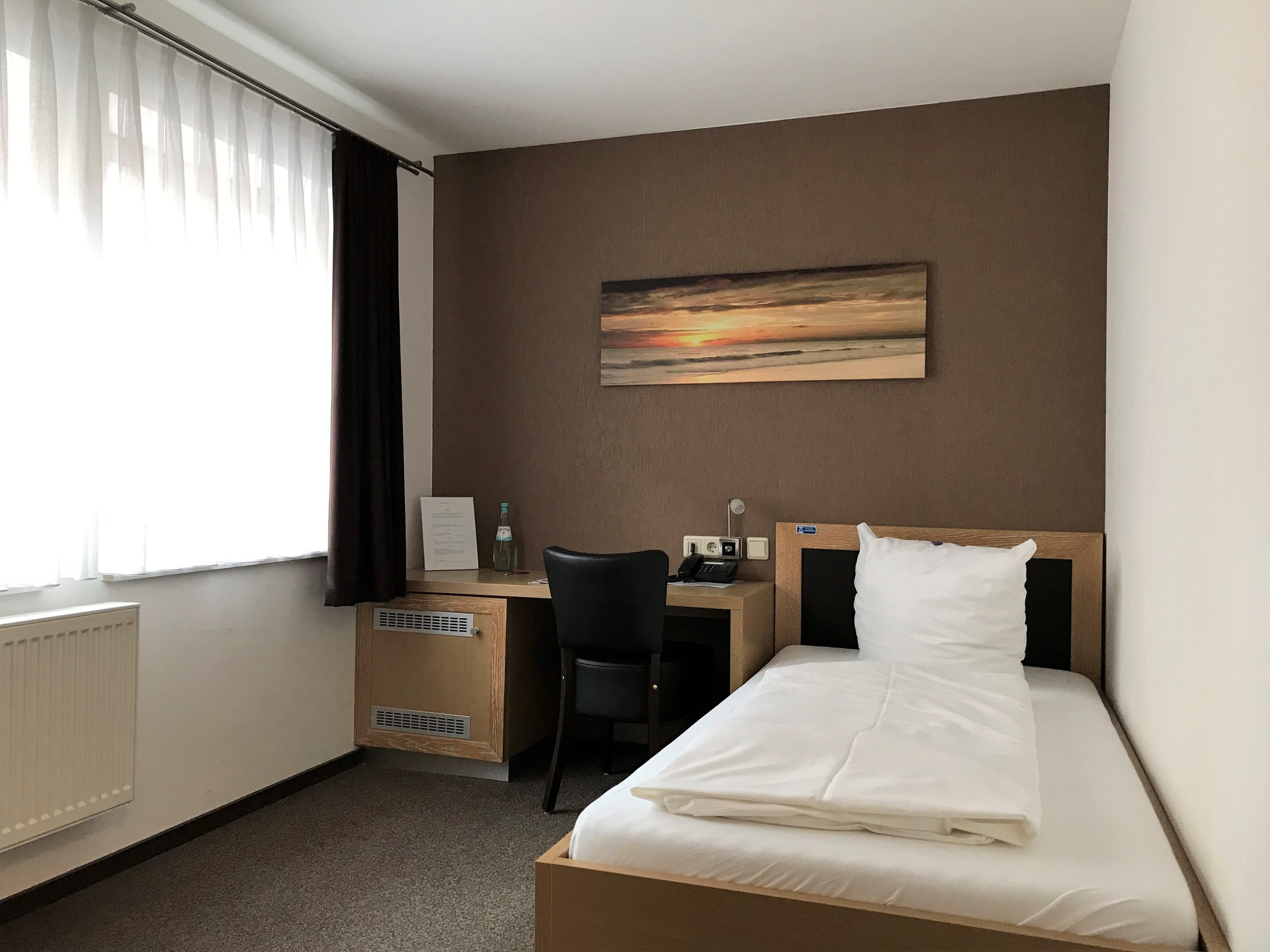 A hotel room with a bed, a desk, a chair, a radiator, a window with curtains, and a painting on the wall.